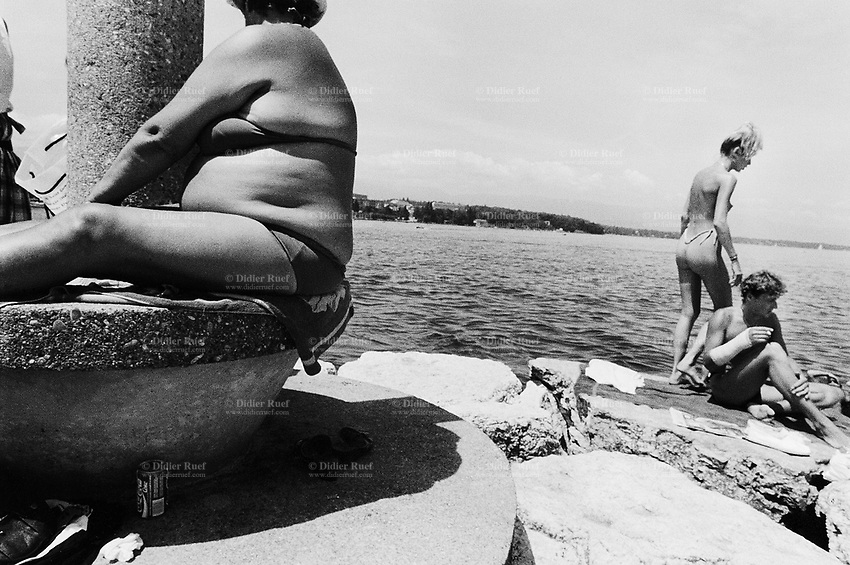 Switzerland. Geneva. Summer on the lake Geneva. A fat woman seats on a concrete platform close to a column. A couple enjoys sunbathing on the rocks. The pretty young woman stands up, wears a string and is topless. The man is seated, smokes a cigarette and has one's fore arm in plaster. © 1987 Didier Ruef
