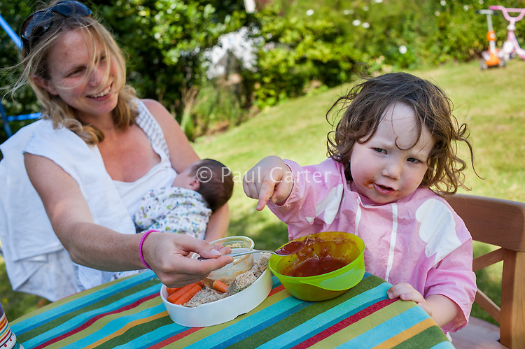 A young mother breastfeeds her new baby whilst feeding her toddler with a spoon at the same time at a table in the garden on a summer's day.<br />
<br />
26/08/12<br />
Hampshire, England, UK