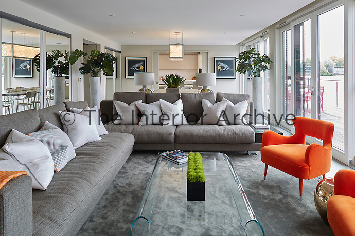 The open plan dining and living space provides ample space for the family to gather and the sliding glass doors lead straight to the terrace. A mirrored wall to one side gives the impression of even more space. A console table with porta roman table lamps define the sitting area which consists of two large grey family sofas and a glass coffee table that sit on an area rug. A pair of orange chairs punctuate the sitting area and add colour and fun to the space.