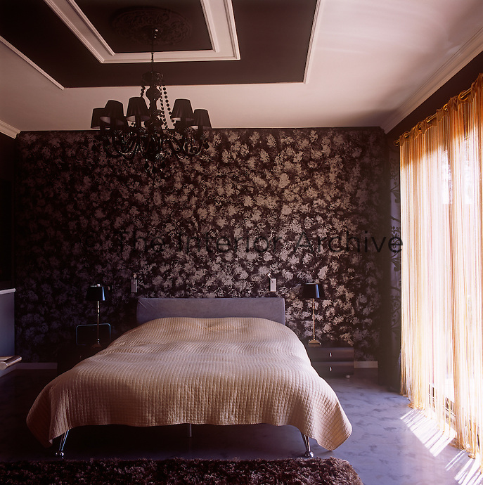 A brown and neutral bedroom with a patterned wallpaper on one wall. A chandelier with brown shades hangs above a double bed with a neutral cover.