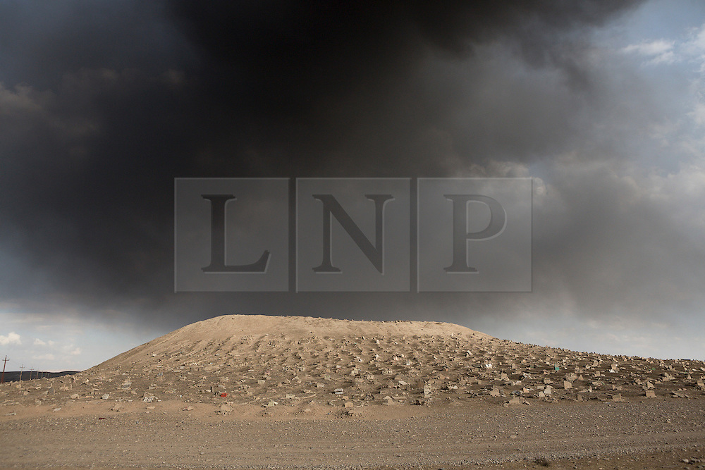 Licensed to London News Pictures. 02/11/2016. Qayyarah, Iraq. Smoke from burning oil wells, set alight by retreating Islamic State militants, clogs the sky over the Qayyarah Cemetery, in the Iraqi town of the same name. Both Shia and Sunni graves within the cemetery were smashed during the town's two year occupation by ISIS extremists who believe that graves should be flat to the earth with no headstone. <br />
<br />
Two months after being liberated from the Islamic State, the Iraqi town of Qayyarah, located around 30km south of Mosul, is still dealing with the environmental repercussions of their ISIS occupation. The town's estimated 15,000 inhabitants constantly live under, and in, heavy clouds of smoke which often envelope the settlement. The clouds emanate from burning oil wells in a nearby oil field that were set alight by retreating ISIS extremists after a two year occupation. The proximity of the fires, often right next to homes within the town, covers many buildings and residents with thick soot and will lead to long term health and environmental implications. Photo credit: Matt Cetti-Roberts/LNP
